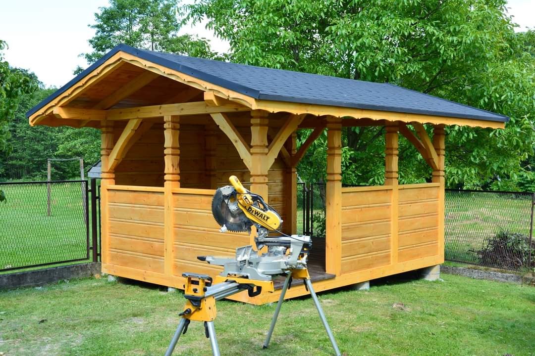 Świeżo wybudowana, jasna, drewniana altana z czarnym, skośnym dachem i dekoracyjnymi kolumnami, na tle zielonego ogrodu; na pierwszym planie ukośnica DeWalt na statywie.