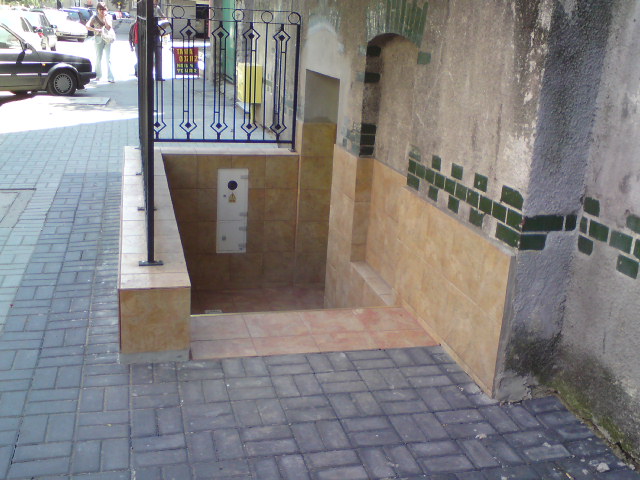 Wejście do piwnicy wyłożone płytkami ceramicznymi, z metalową balustradą i skrzynką elektryczną na ścianie, widoczne z poziomu chodnika.