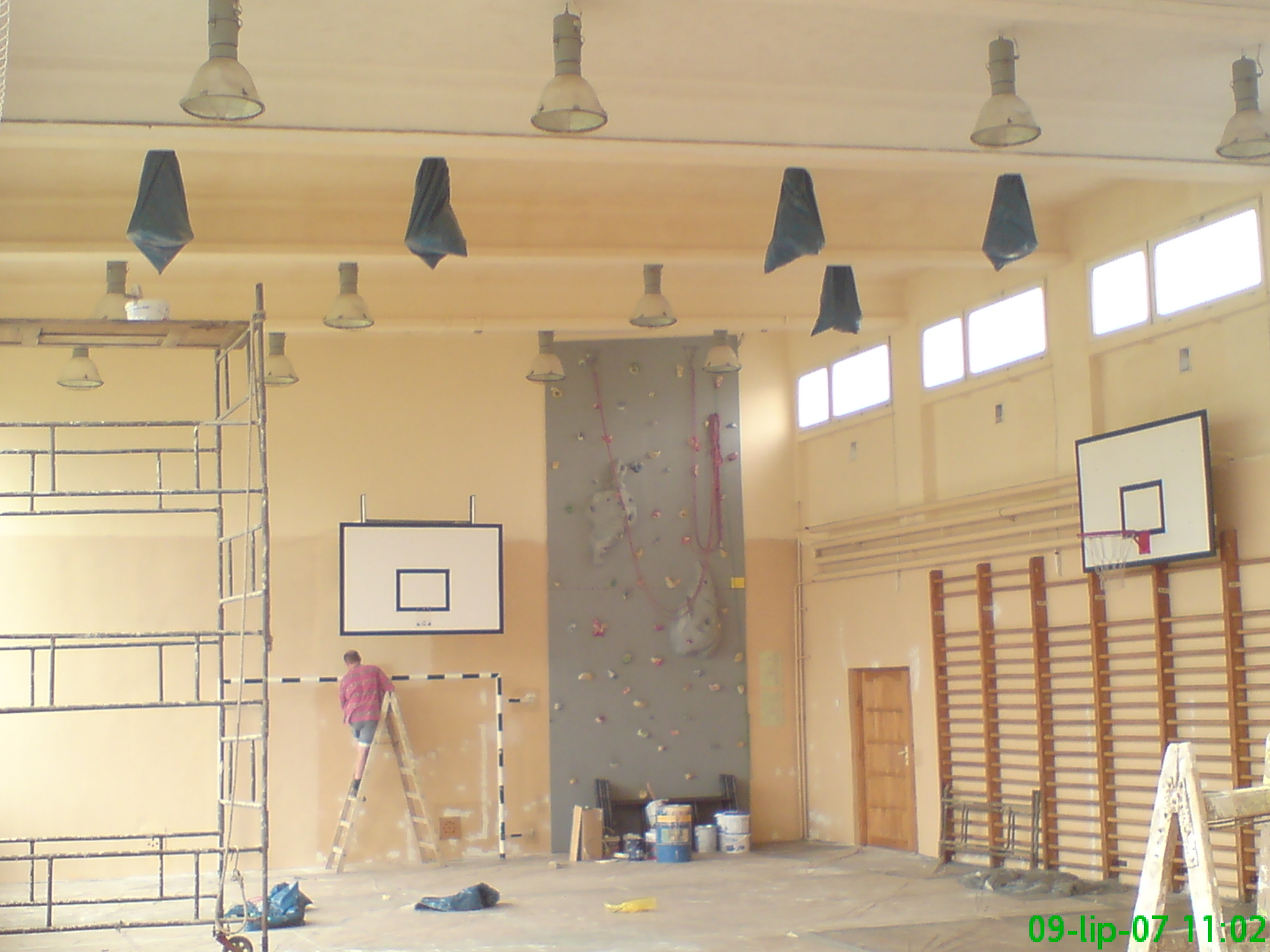 Malowanie ściany w sali gimnastycznej z drabinkami, koszem i ścianką wspinaczkową, widoczne rusztowanie i wiadro z farbą.