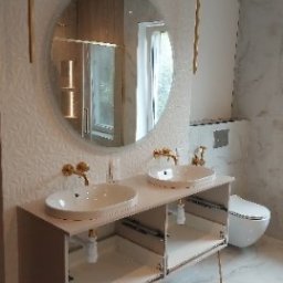 MAGRA Izabella Kalinowska - Nowoczesna łazienka z podwójną umywalką nablatową na szafce z otwartymi półkami, okrągłe lustro i złote akcenty armatury oraz lamp wiszących.