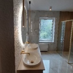 MAGRA Izabella Kalinowska - Wykończona łazienka z podwójną umywalką nablatową, złotą armaturą i oświetleniem LED w okrągłym lustrze. Widoczna kabina prysznicowa ze złotymi profilami i marmurowe płytki na podłodze i ścianach.
