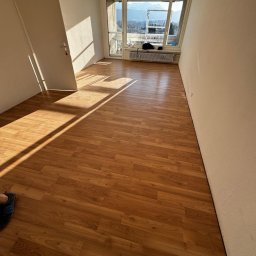 MAGRA Izabella Kalinowska - Puste pomieszczenie z nowo ułożonymi panelami podłogowymi w odcieniach brązu, widoczne światło słoneczne wpadające przez okno balkonowe z widokiem na góry.