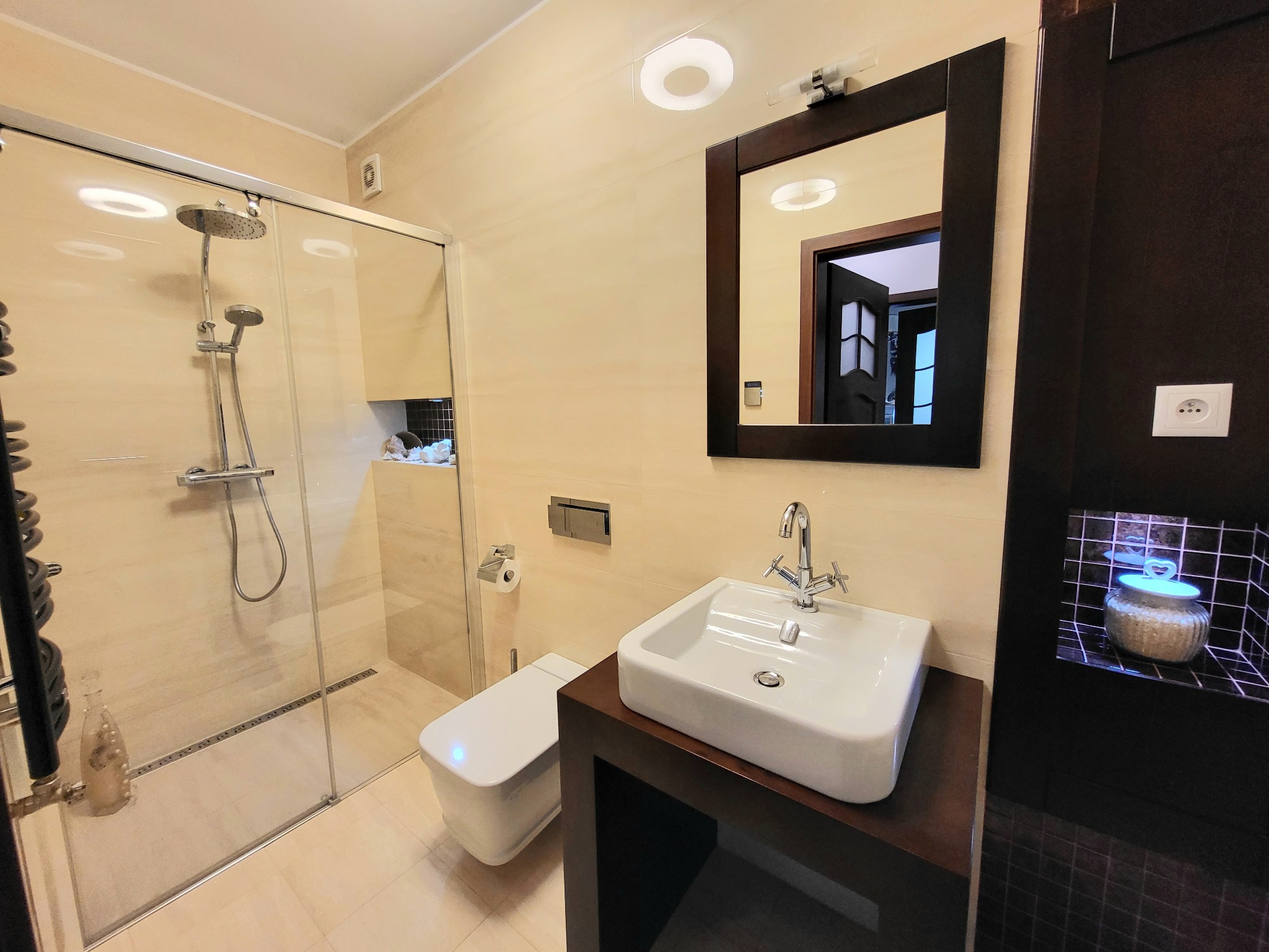 Wykończona łazienka w beżowej kolorystyce z kabiną prysznicową typu walk-in, umywalką nablatową na ciemnobrązowej szafce i lustrem w ciemnej ramie. W tle widoczna toaleta i dekoracyjna wnęka...