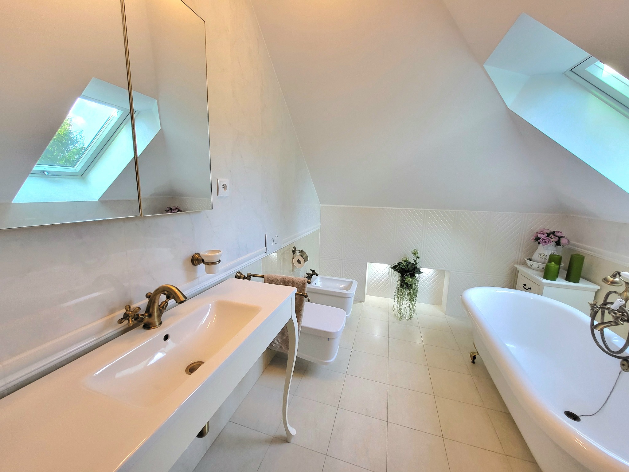 Jasna łazienka na poddaszu z oknem dachowym, białą wanną na lwich nóżkach, umywalką z baterią w stylu retro i dekoracyjną ścianą z białych płytek.