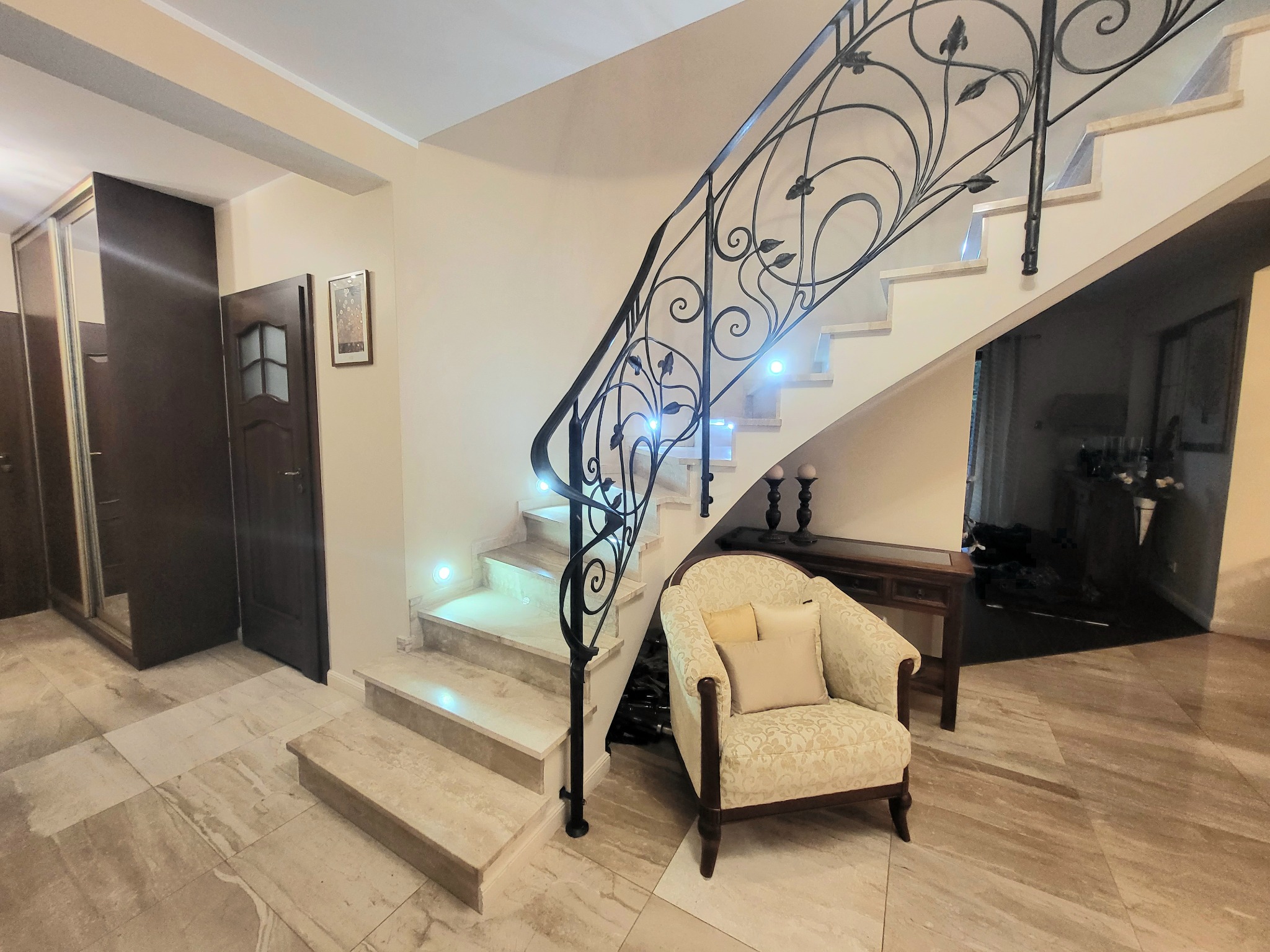 Eleganckie wnętrze: marmurowe schody z kutą, ozdobną balustradą, podświetlane stopnie, fotel w kwiatowy wzór z poduszkami, marmurowa podłoga i drewniane drzwi.