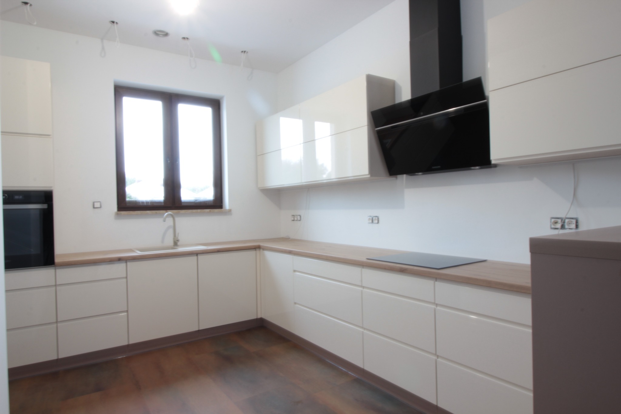 Jasna, nowoczesna kuchnia z białymi szafkami, drewnianym blatem i ciemnobrązową podłogą, widok na okno i czarny okap