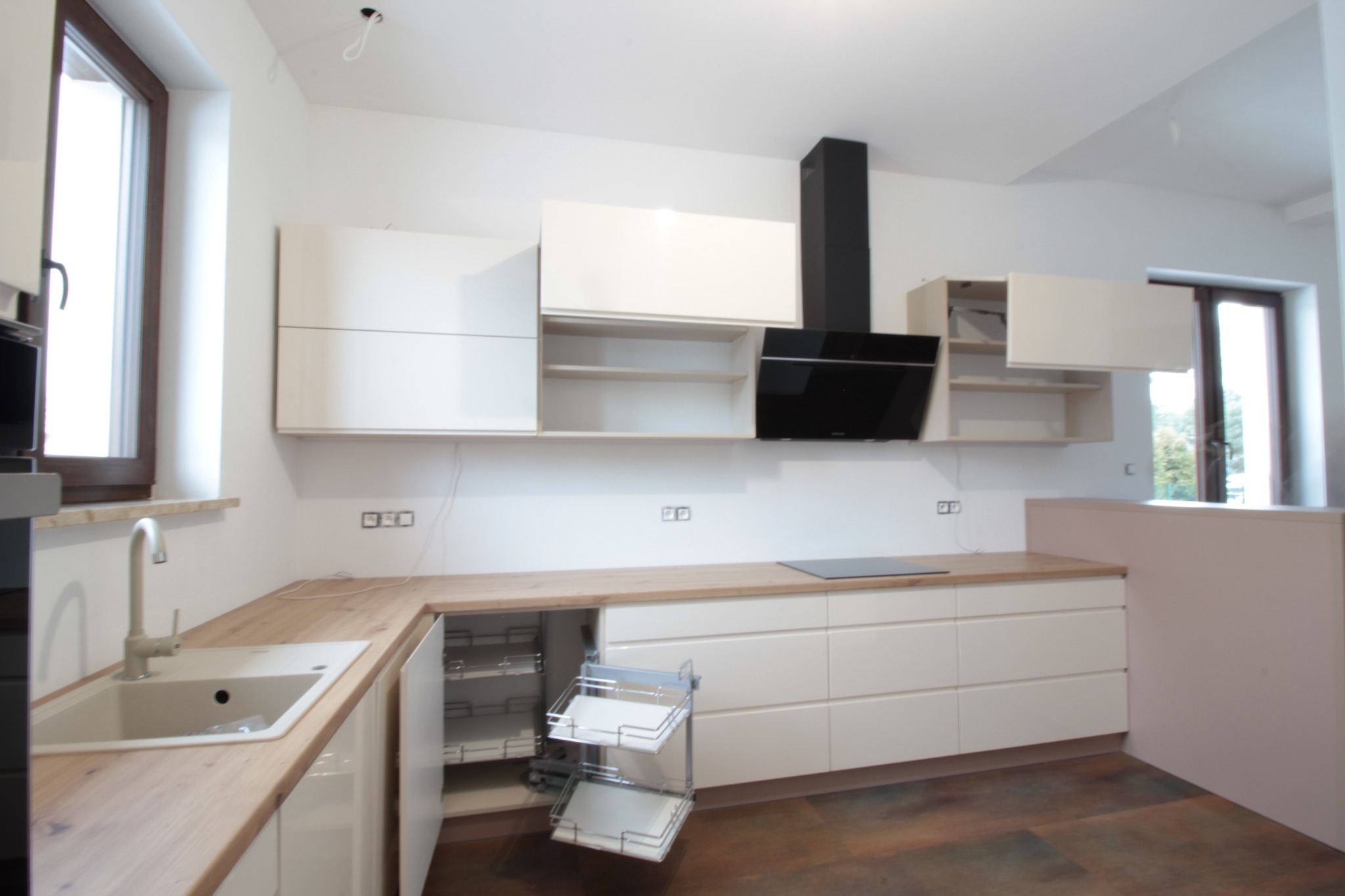 Nowoczesna, jasna kuchnia z białymi szafkami i drewnianym blatem, widoczny wysuwany system koszy cargo w szafce dolnej, czarny okap nad płytą grzewczą.