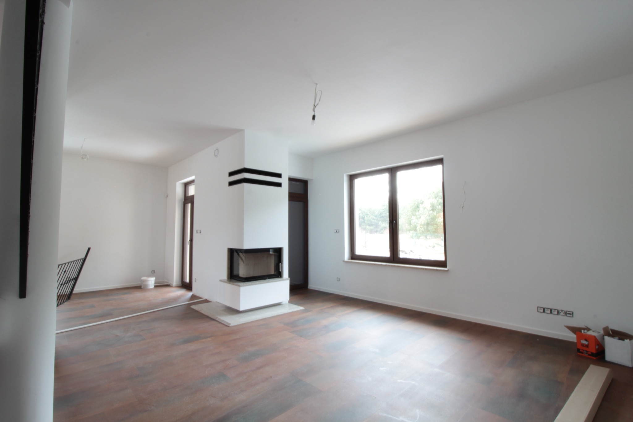 Jasne, przestronne wnętrze w trakcie prac wykończeniowych, z ciemnobrązową podłogą imitującą drewno, białymi ścianami, nowoczesnym kominkiem z czarnymi akcentami i dużym oknem z widokiem na zieleń...