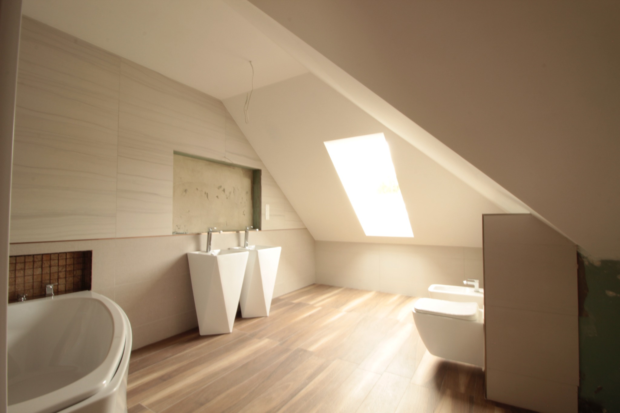 Jasne poddasze łazienki z drewnopodobną podłogą, białą wanną, dwoma umywalkami na postumentach i toaletą, częściowo wykończone płytki na ścianach, wnęka z mozaiką i niedokończone fragmenty ścian.