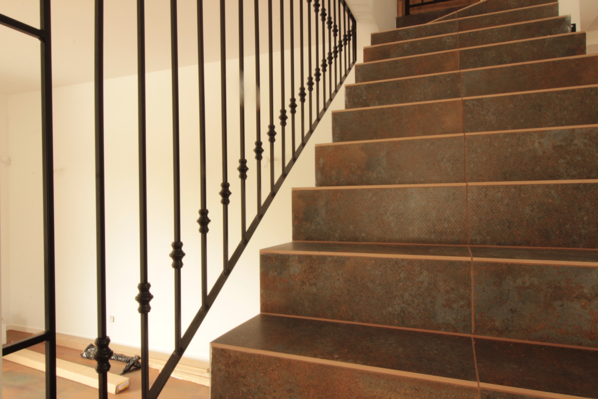 Schody wyłożone płytkami imitującymi rdzę, z czarną, kutą balustradą, widok z dołu, materiały budowlane i narzędzia na podłodze w tle.