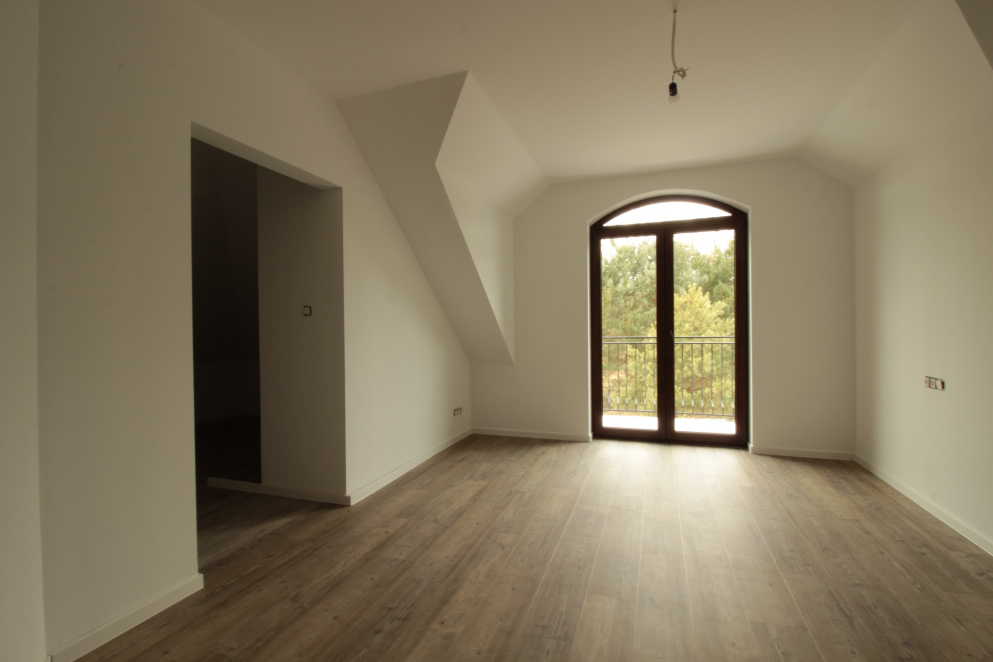 Puste, jasne poddasze z drewnianą podłogą, oknem balkonowym z widokiem na zieleń i otwartym przejściem do innego pomieszczenia, widoczne gniazdka elektryczne i kabel od lampy wiszącej.