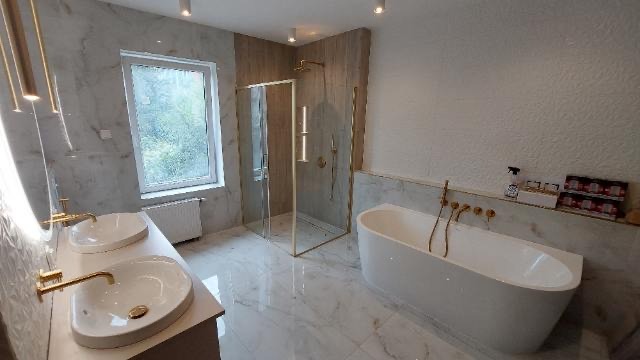 Luksusowa łazienka z marmurowymi ścianami i podłogą, kabina prysznicowa ze złotymi detalami, wolnostojąca wanna z baterią ścienną, dwie umywalki nablatowe z armaturą w kolorze złota, oświetlenie...