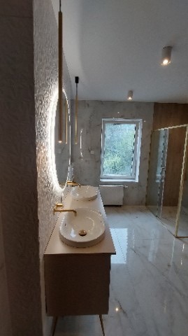 Wykończona łazienka z podwójną umywalką nablatową, złotą armaturą i oświetleniem LED w okrągłym lustrze. Widoczna kabina prysznicowa ze złotymi profilami i marmurowe płytki na podłodze i ścianach.