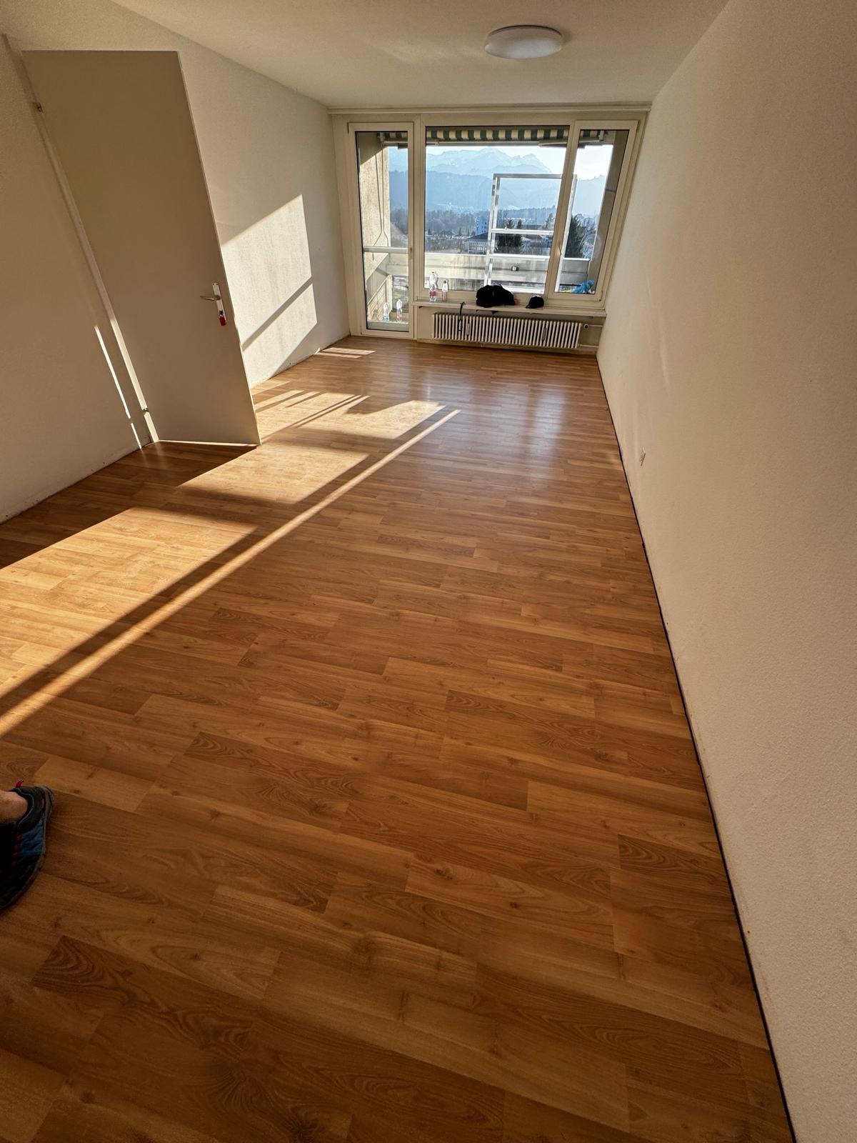 Puste pomieszczenie z nowo ułożonymi panelami podłogowymi w odcieniach brązu, widoczne światło słoneczne wpadające przez okno balkonowe z widokiem na góry.