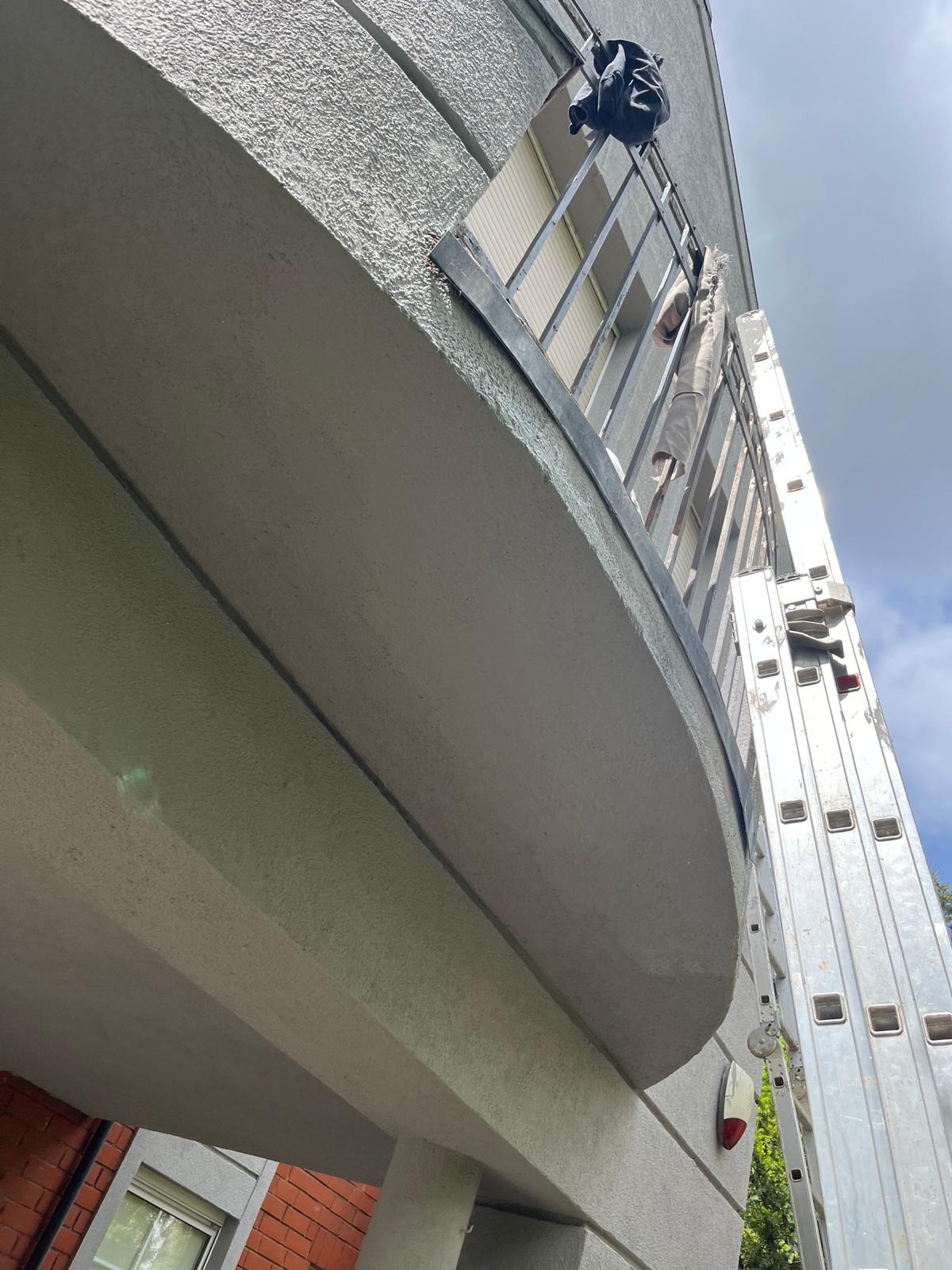 Prace na balkonie z drabiny aluminiowej; widoczny fragment balkonu z szarą elewacją, na balustradzie wisi plecak i ubranie, drabina oparta o balkon.