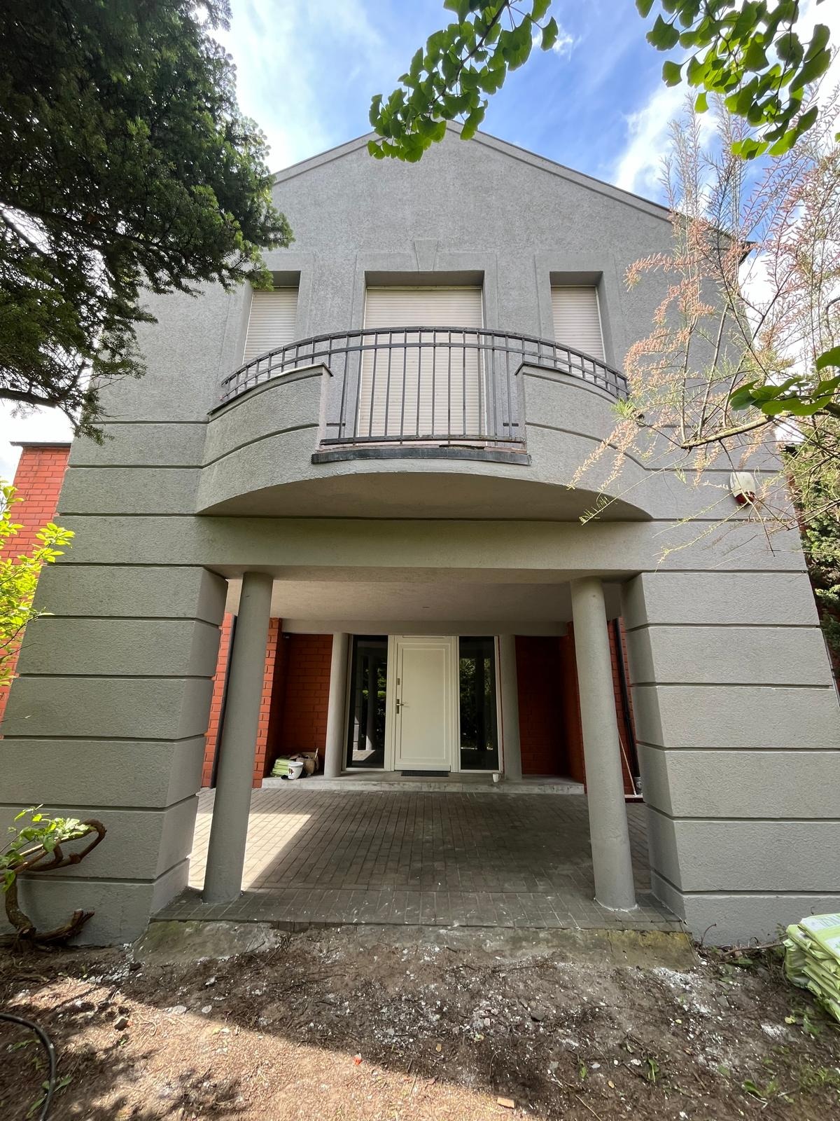 Szary, dwupiętrowy dom z balkonem, widok z dołu, z kolumnami podtrzymującymi zadaszenie nad wejściem, widoczne worki z materiałami budowlanymi obok wejścia.