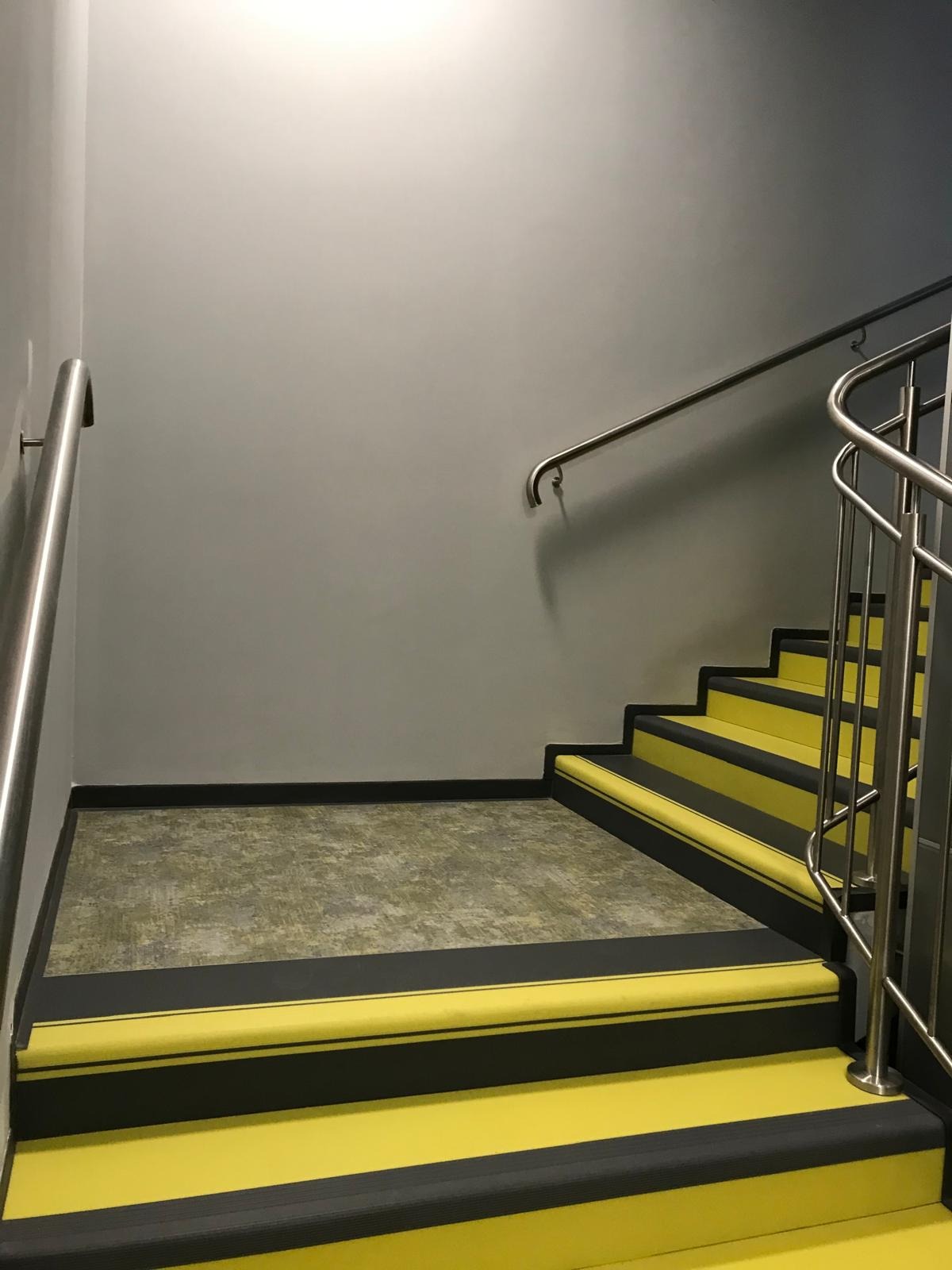 Schody wewnętrzne z żółtymi stopniami i czarnymi krawędziami, zabezpieczone metalową barierką, widok z dołu.