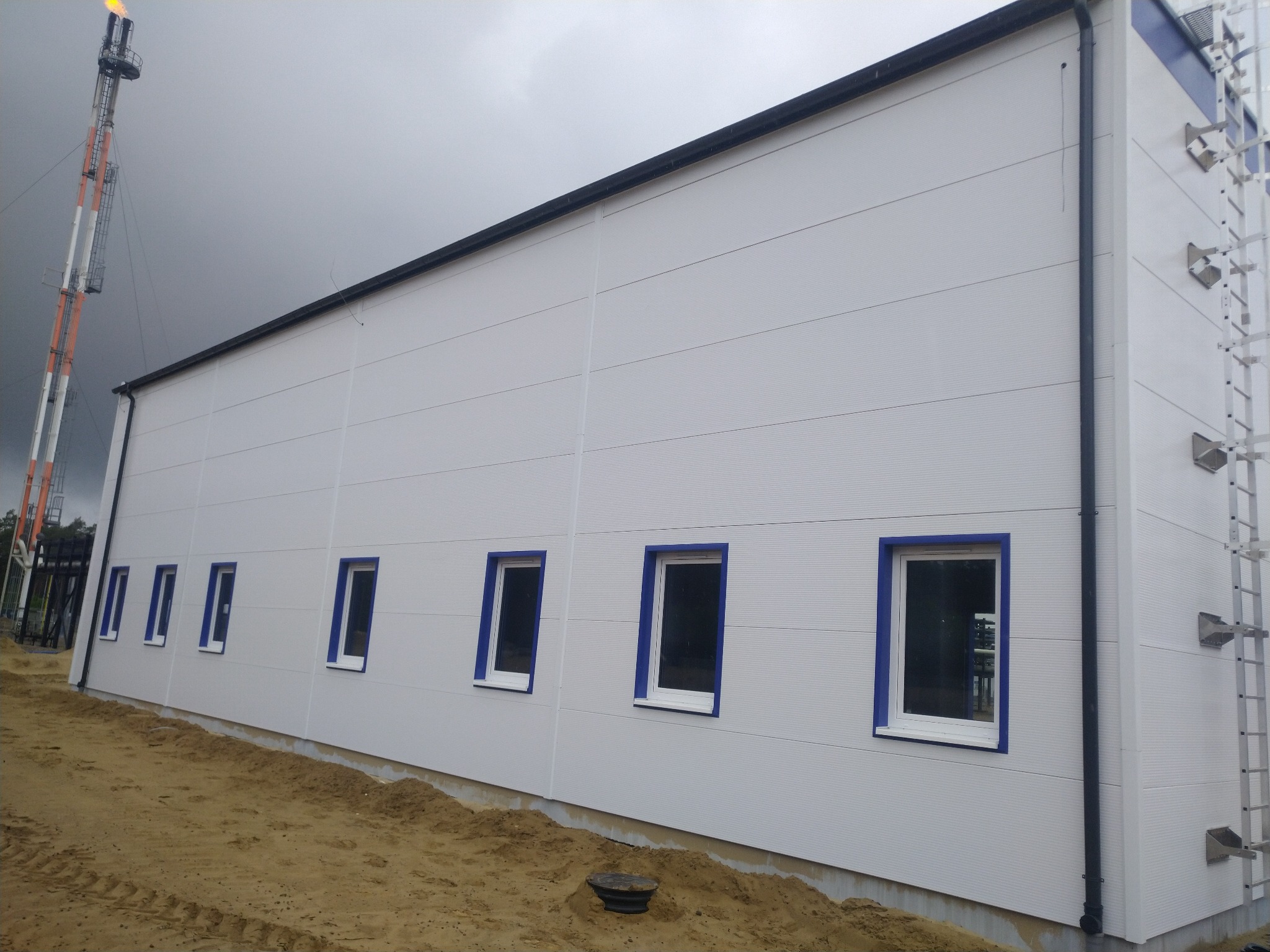 Boczny widok hali przemysłowej z białych płyt warstwowych z rzędem okien w niebieskich ramach i zewnętrzną drabiną po prawej stronie, na zewnątrz widoczna instalacja przemysłowa z płomieniem.