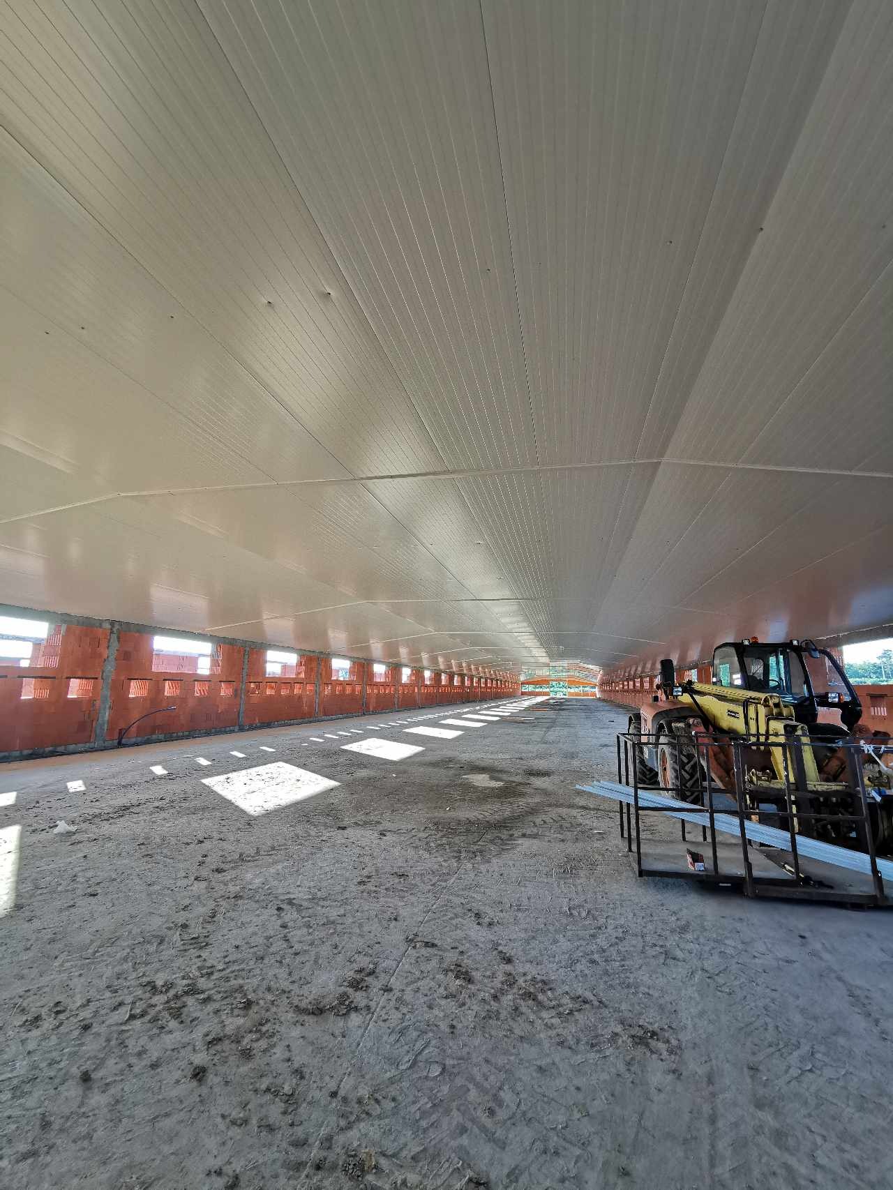 Wnętrze hali w trakcie budowy, widoczne ściany z pustaków, zamontowane płyty warstwowe na suficie oraz żółta ładowarka teleskopowa z paletą profili.