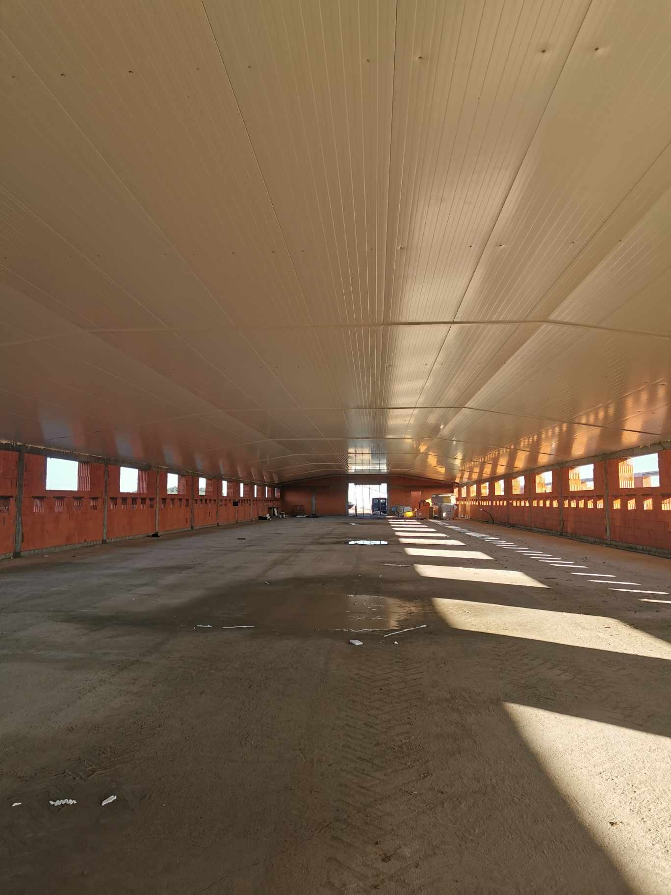 Wnętrze nowo budowanej hali z widoczną konstrukcją ścian z pustaków oraz zamontowanym sufitem z płyt warstwowych, na podłodze widoczne ślady opon i kałuże.