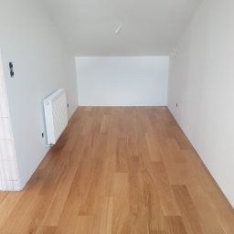 Studio Podłóg Natur Wood - Jasna drewniana podłoga w nowym, pomalowanym na biało pokoju z grzejnikiem na ścianie i widocznymi przewodami elektrycznymi na suficie.
