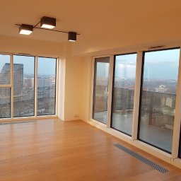 Studio Podłóg Natur Wood - Jasne, przestronne wnętrze apartamentu z nową, gładką, drewnianą podłogą w odcieniach brązu, dużymi oknami z widokiem na miasto i nowoczesnym oświetleniem sufitowym.