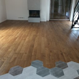 Studio Podłóg Natur Wood - Jasna, drewniana podłoga ułożona we wzór jodełki przechodzi w szare, heksagonalne płytki podłogowe w salonie z kominkiem i czarnymi schodami.