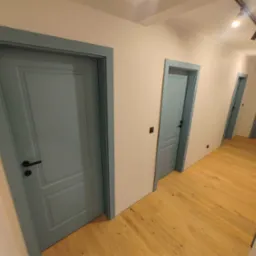 Korytarz z trzema parami drzwi w odcieniach błękitu, drewniana podłoga z wyraźnym usłojeniem, oświetlenie szynowe.