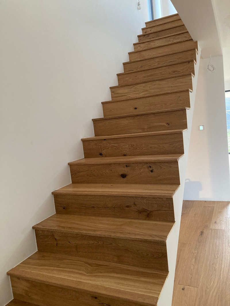 Drewniane schody z widocznymi sękami, prowadzące w górę w jasnym wnętrzu z panelami podłogowymi.