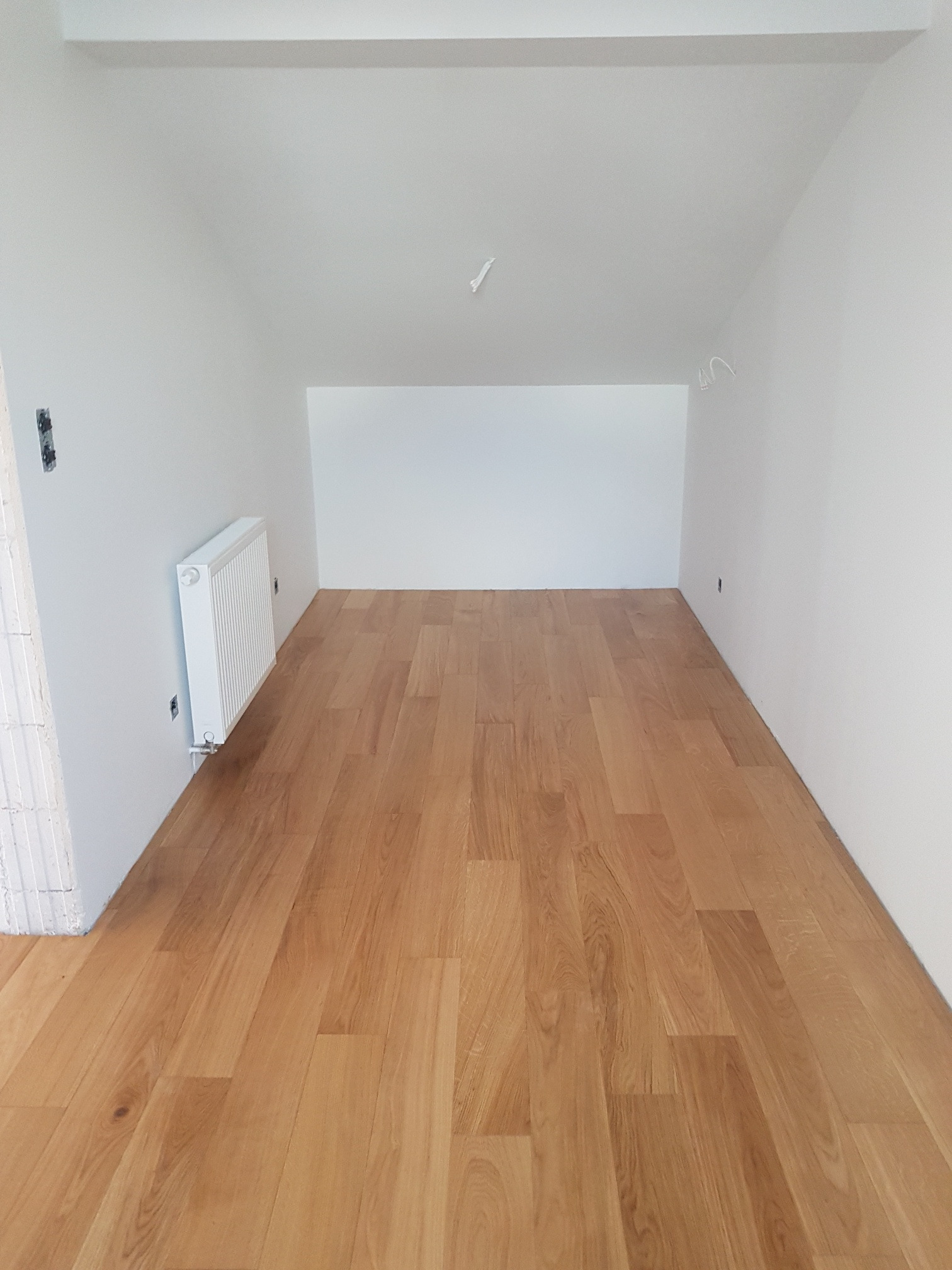 Jasna drewniana podłoga w nowym, pomalowanym na biało pokoju z grzejnikiem na ścianie i widocznymi przewodami elektrycznymi na suficie.