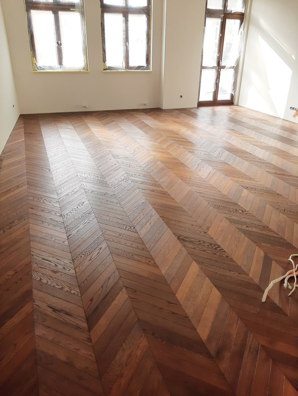 Ukończona podłoga z ciemnego drewna ułożona we wzór jodełki w jasnym pokoju z oknami zabezpieczonymi folią malarską.