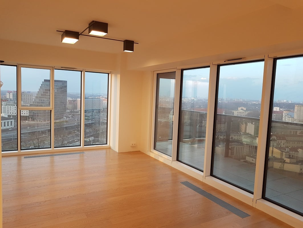 Jasne, przestronne wnętrze apartamentu z nową, gładką, drewnianą podłogą w odcieniach brązu, dużymi oknami z widokiem na miasto i nowoczesnym oświetleniem sufitowym.