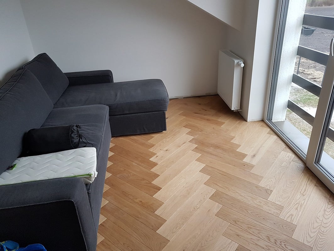 Jasna, dębowa podłoga ułożona w jodełkę francuską, widoczna z perspektywy narożnika pokoju z szarą sofą, białym grzejnikiem i oknem balkonowym.