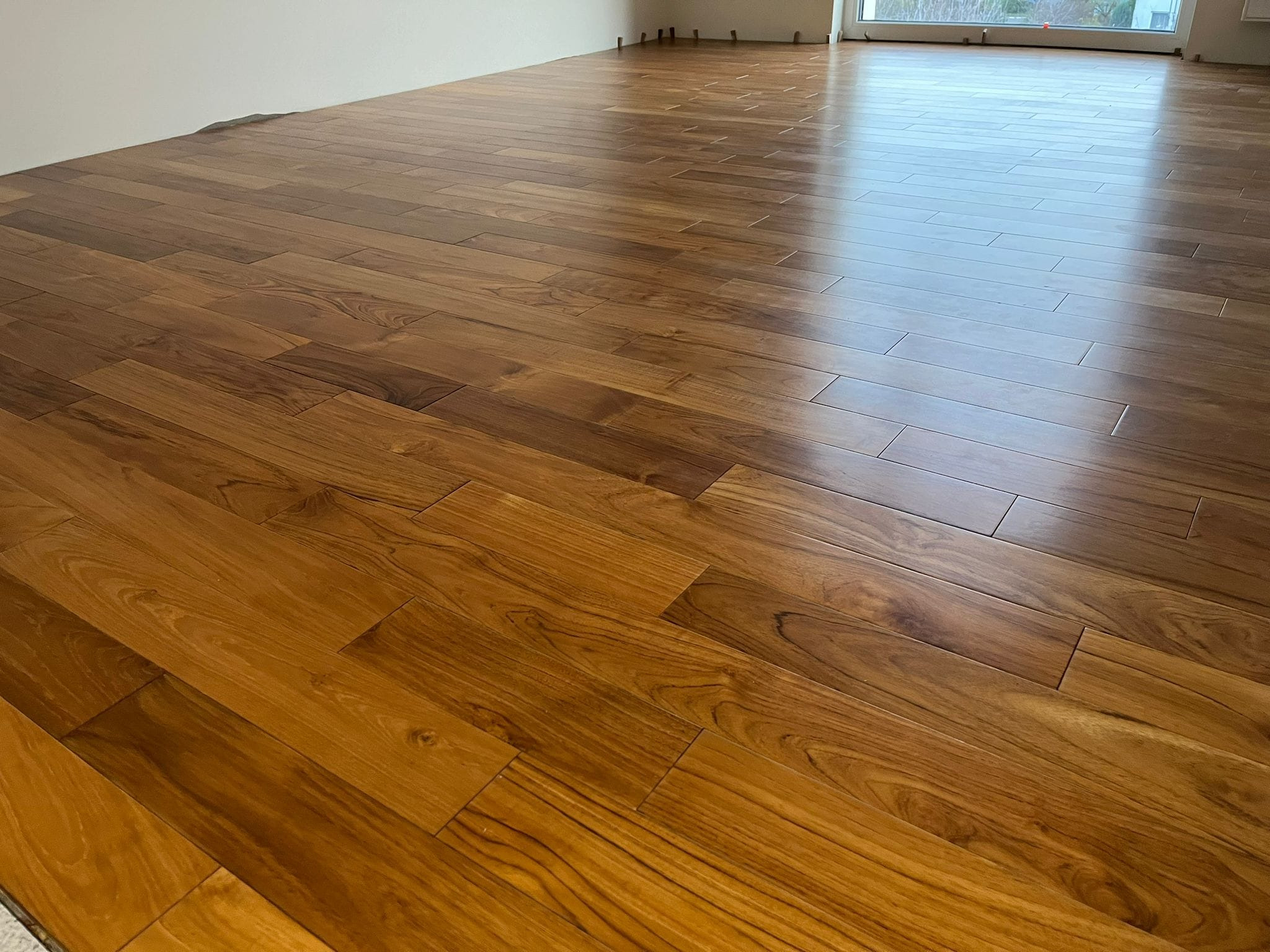 Nowo ułożona, błyszcząca podłoga z egzotycznego drewna, widoczne listwy dystansowe przy ścianie, odbicie światła z okna.