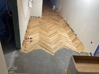 Układanie parkietu w jodełkę francuską w korytarzu, widoczne niedokończone fragmenty i narzędzia montażowe.