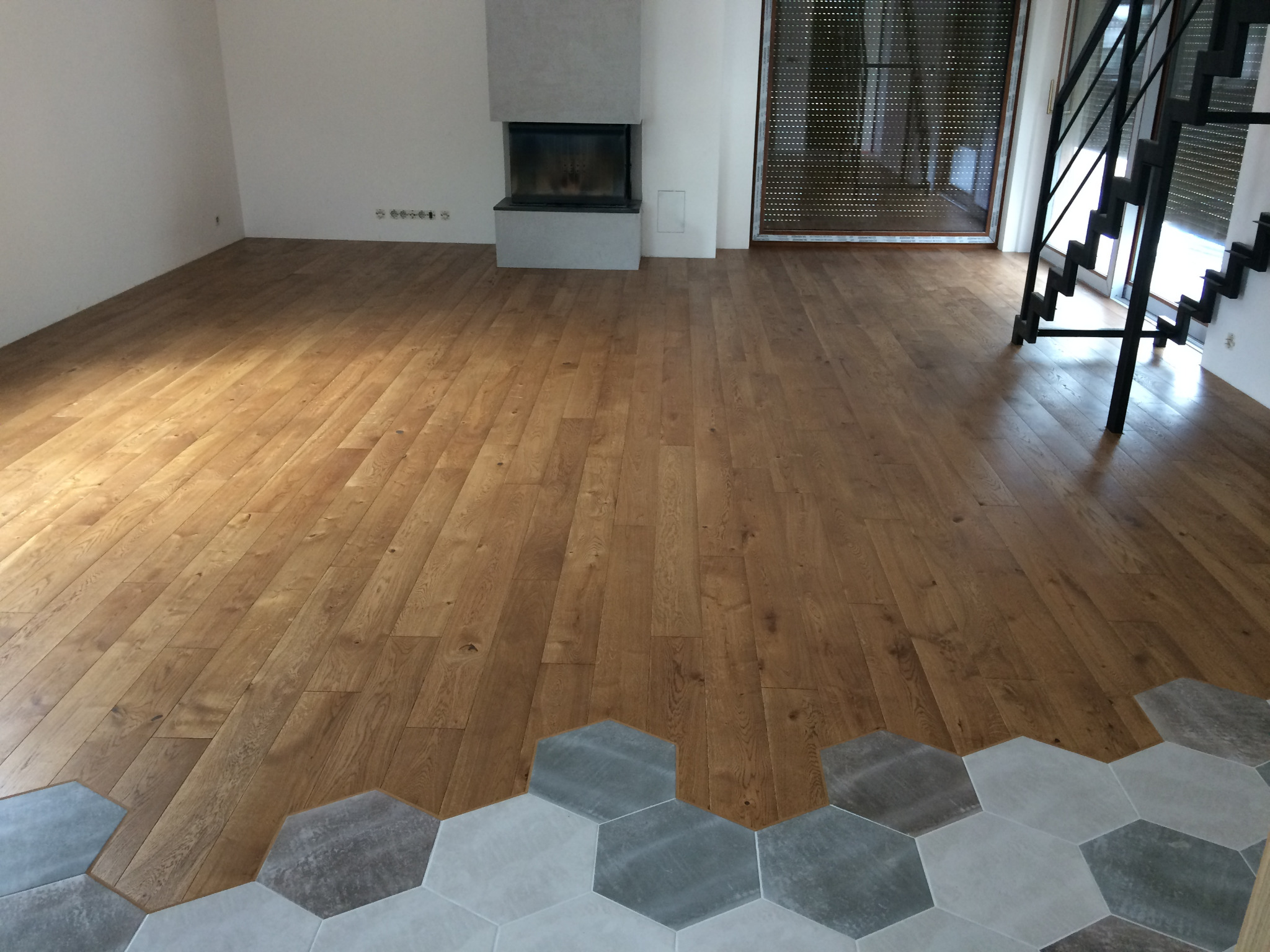 Jasna, drewniana podłoga ułożona we wzór jodełki przechodzi w szare, heksagonalne płytki podłogowe w salonie z kominkiem i czarnymi schodami.