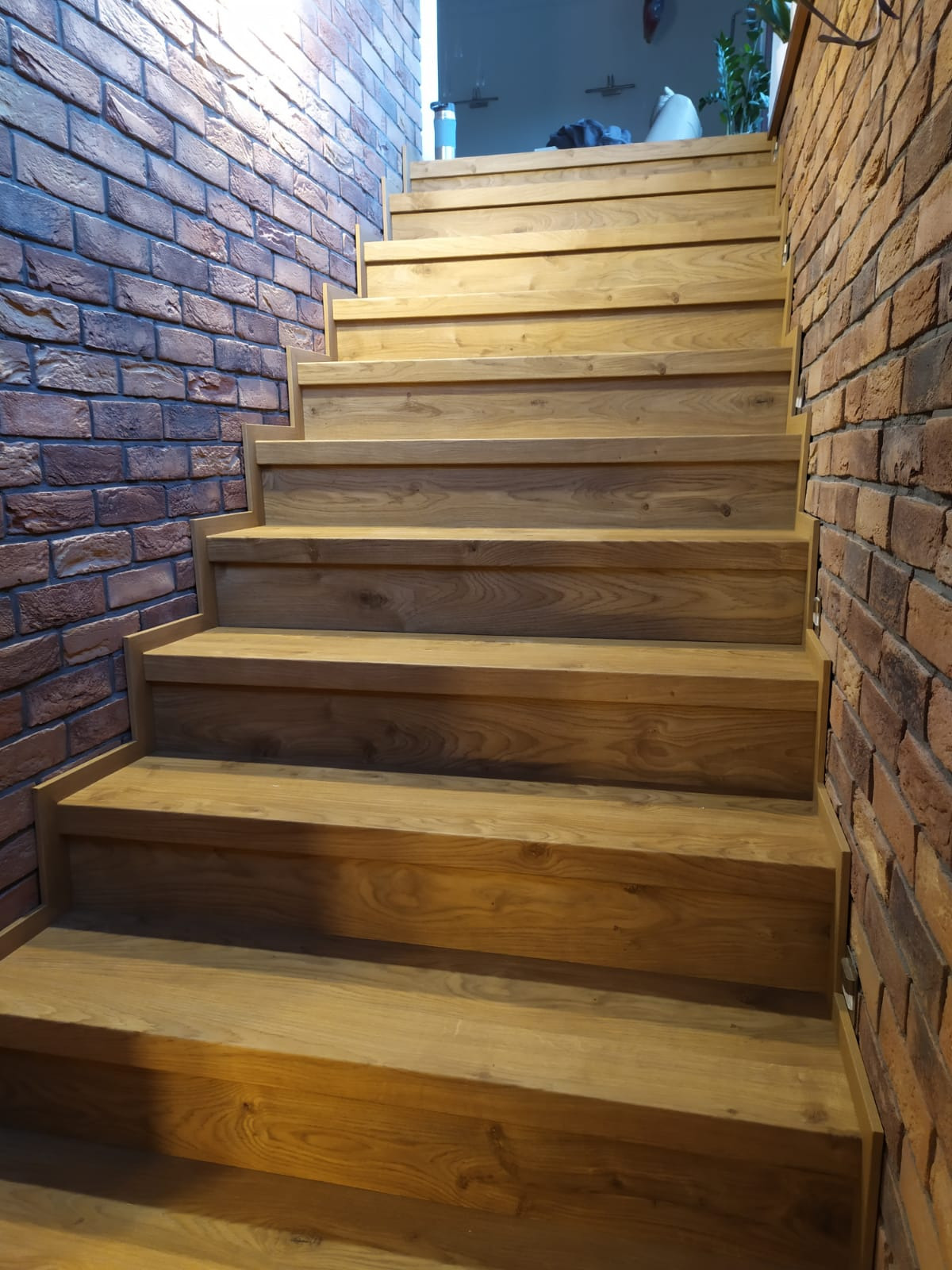 Drewniane schody z dębowymi stopniami, otoczone ścianami z cegły dekoracyjnej w odcieniach brązu i czerwieni, z widocznym przejściem do innego pomieszczenia w tle.