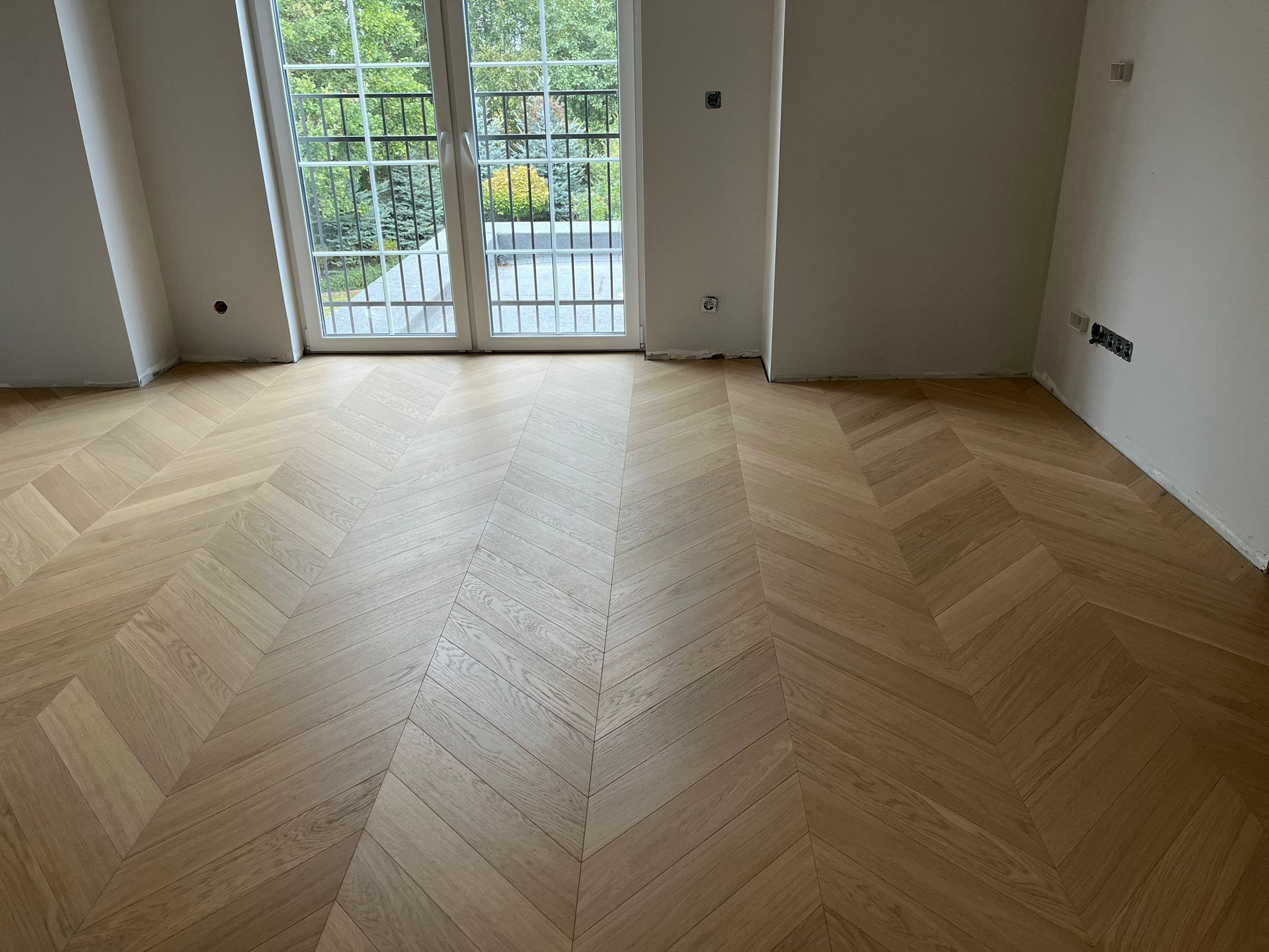 Jasny parkiet ułożony we wzór jodełki francuskiej w nowym pomieszczeniu z białymi ścianami i oknami balkonowymi wychodzącymi na zieleń.