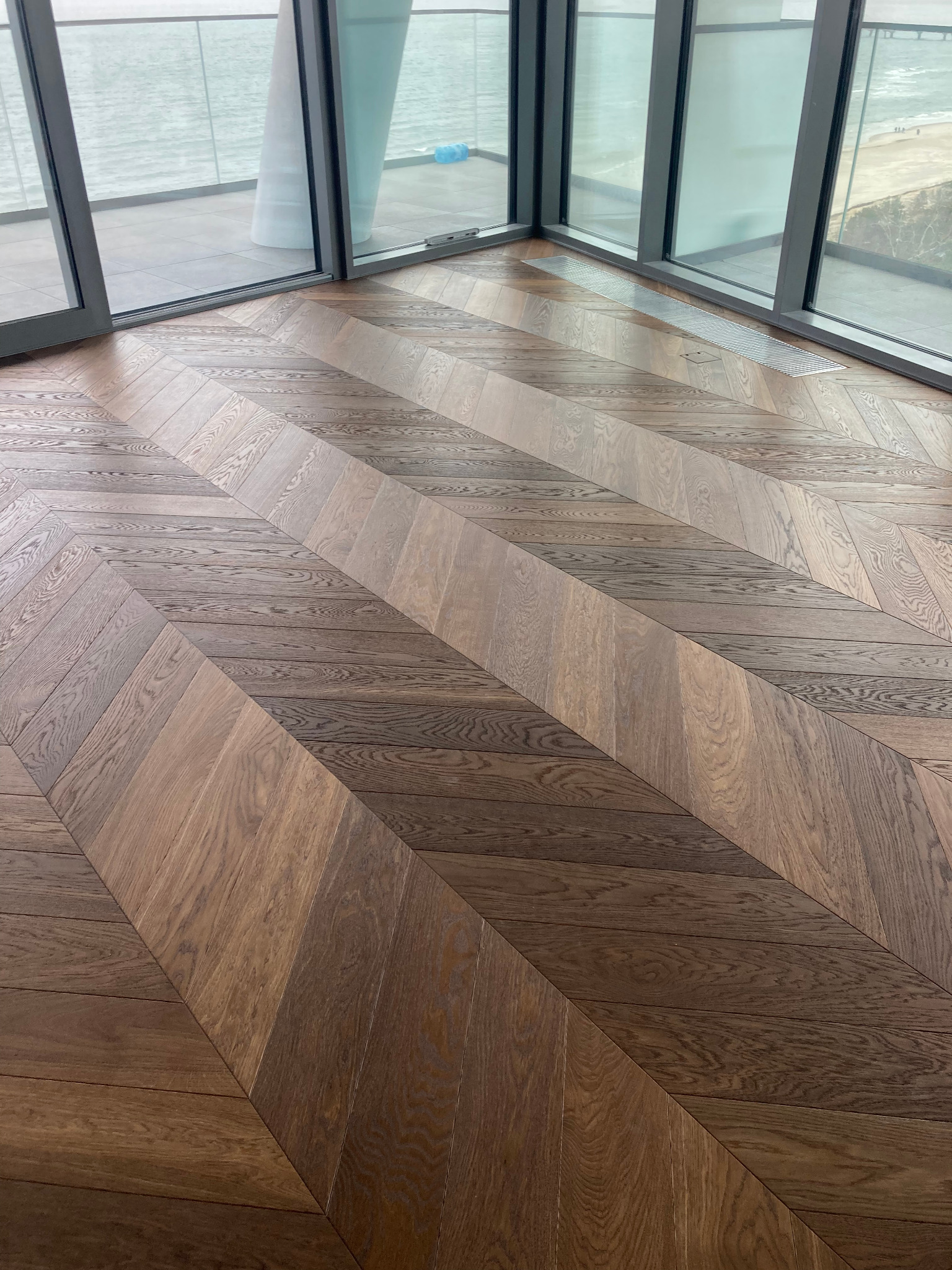 Parkiet ułożony w jodełkę francuską, z widocznym usłojeniem drewna, w nowoczesnym wnętrzu z dużymi oknami i widokiem na morze.