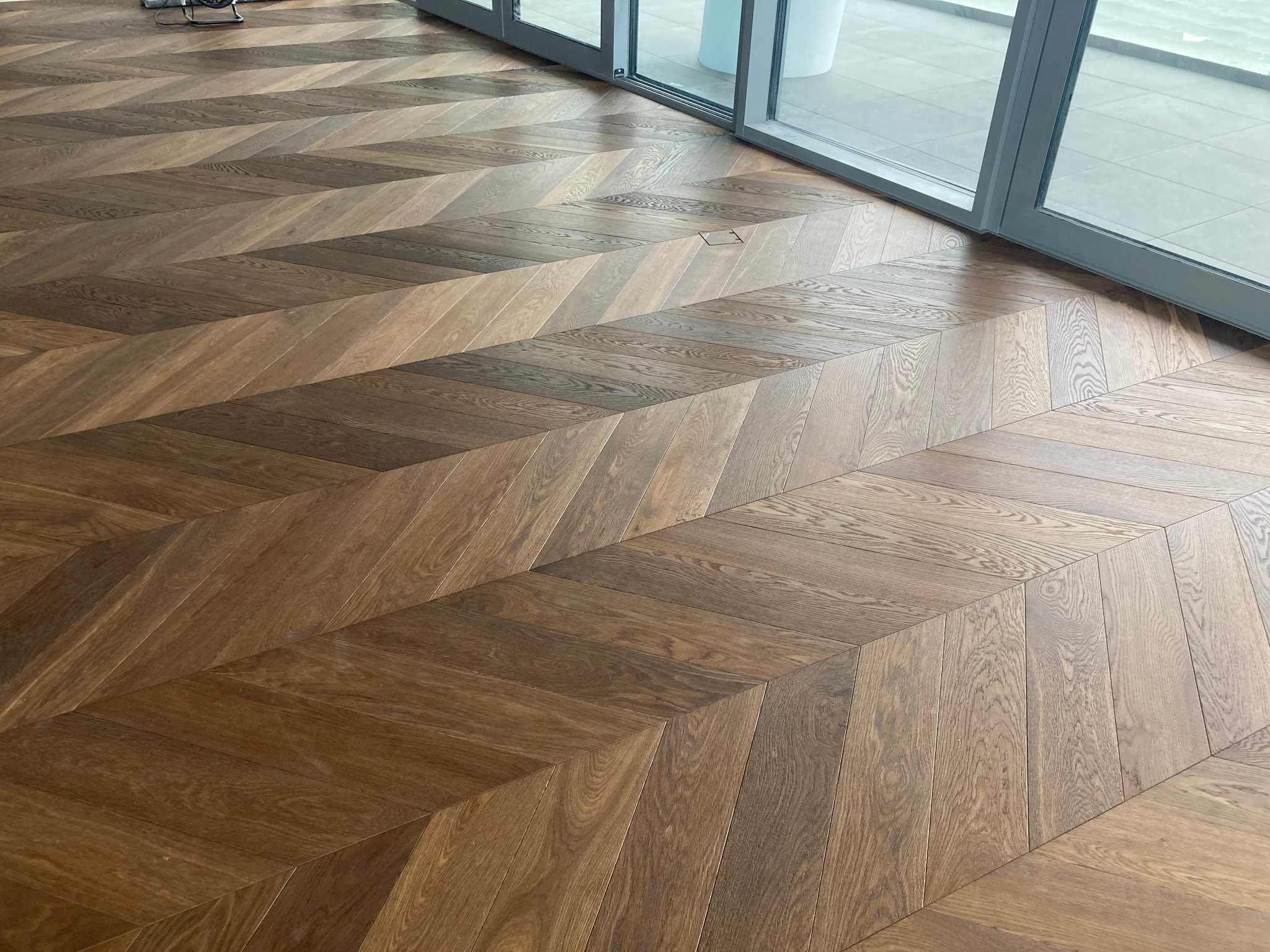 Ułożona w jodełkę, lakierowana podłoga z drewnianych desek w odcieniach brązu, z widocznym fragmentem okna przesuwanego.