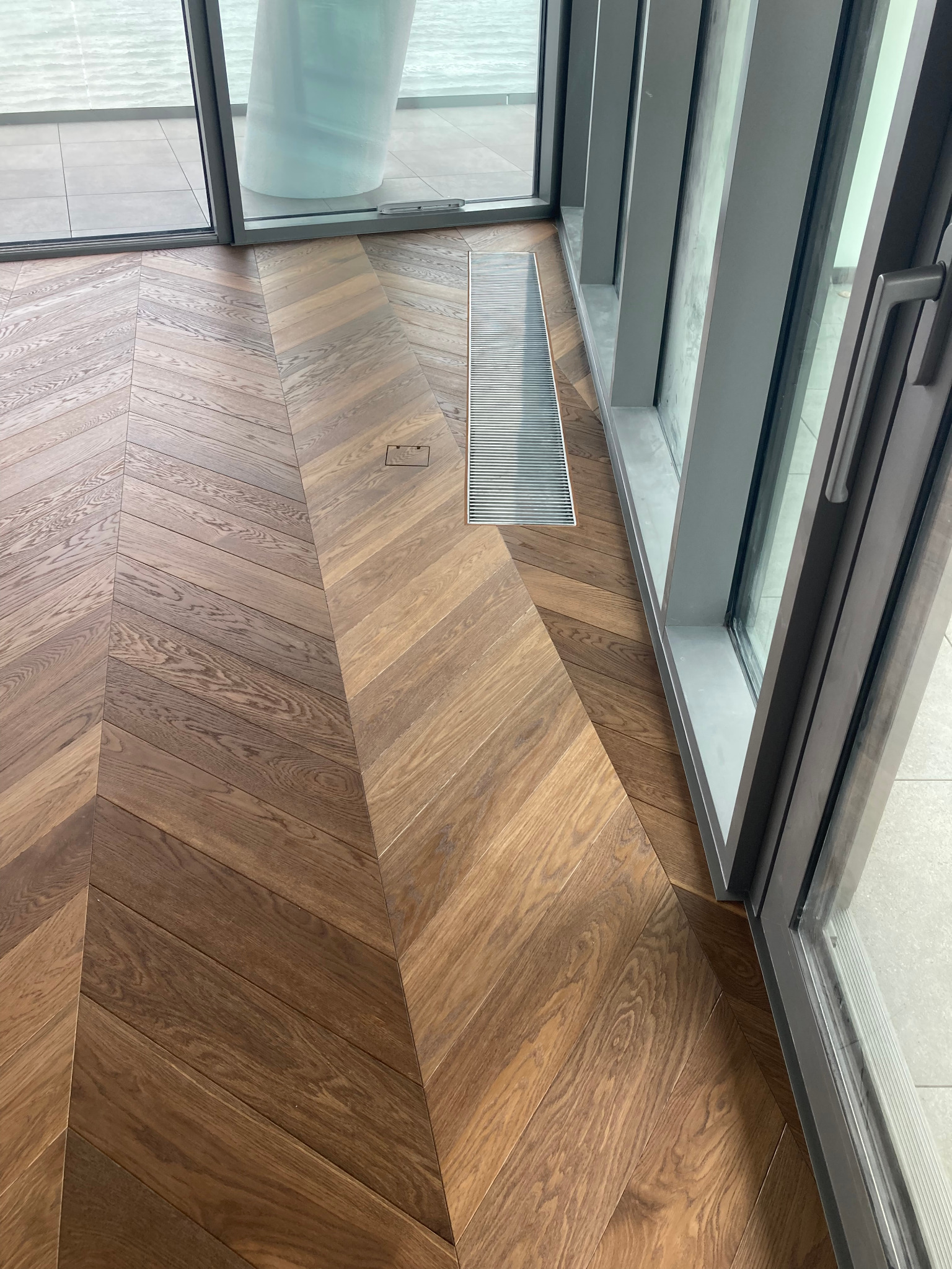 Parkiet ułożony we wzór jodełki francuskiej, z widocznym odpływem liniowym przy oknie, oświetlonym naturalnym światłem.