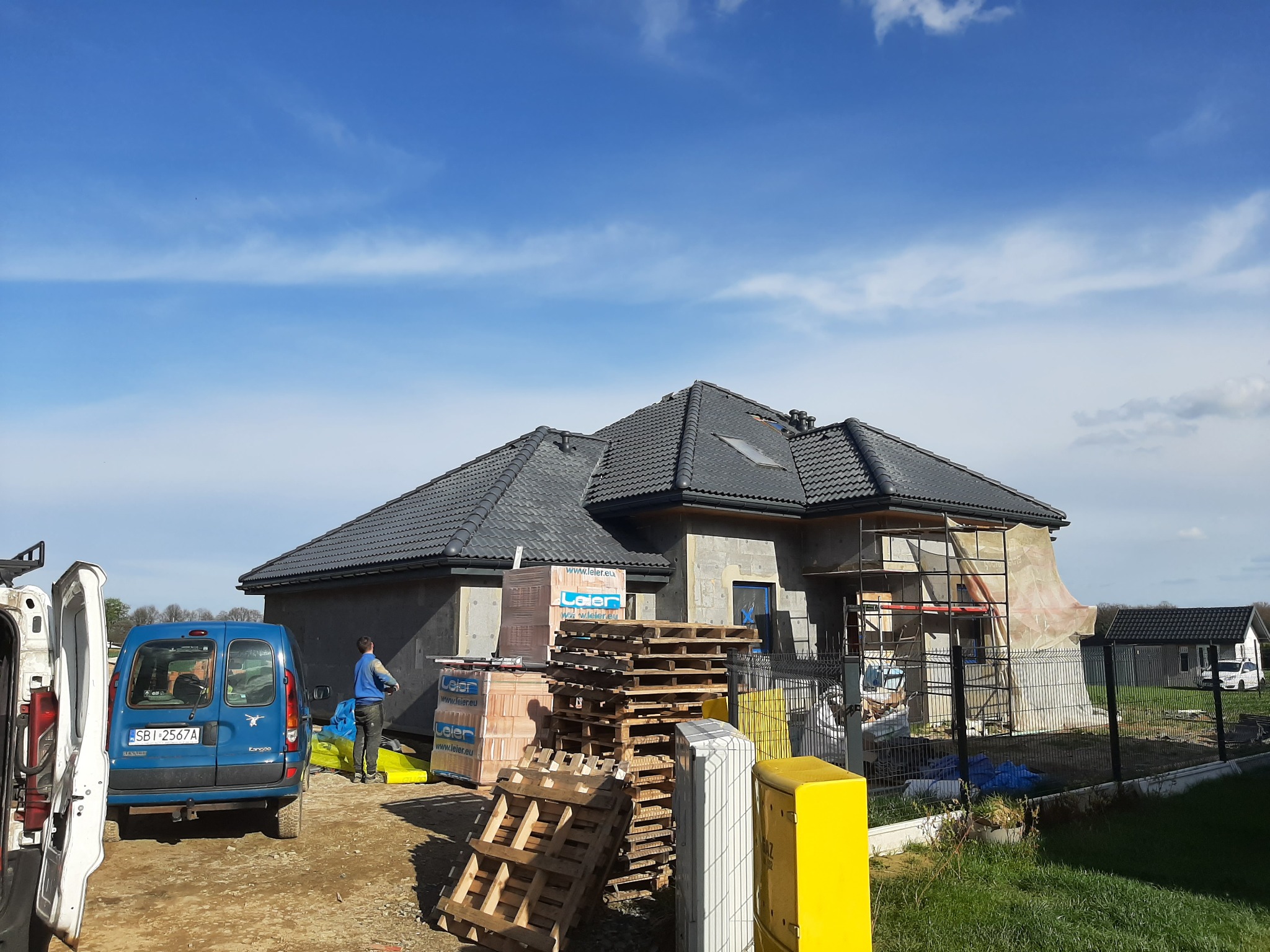 Budowa domu jednorodzinnego z ciemną dachówką karpiówką, widoczne palety z materiałami budowlanymi i rusztowania.