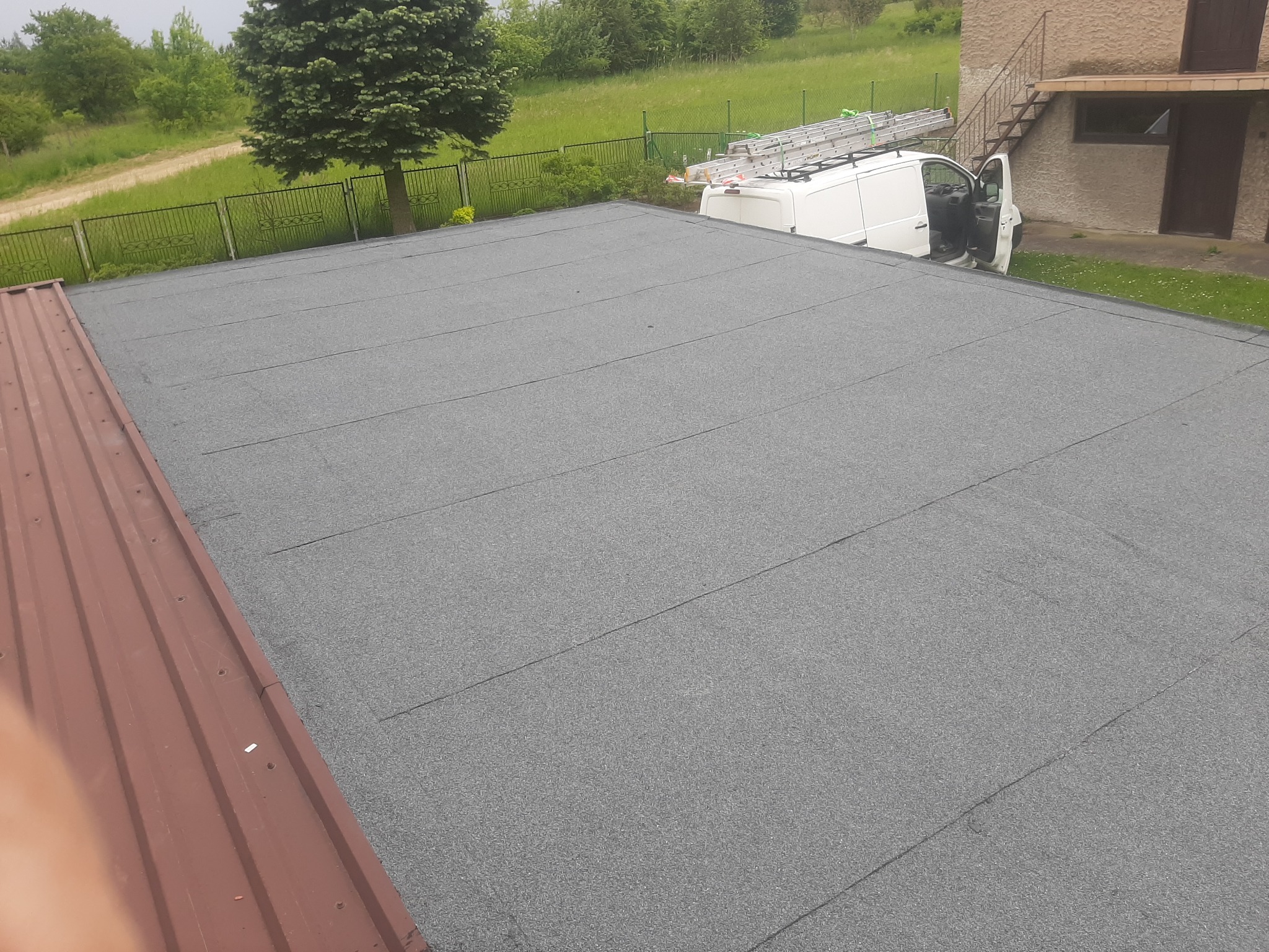 Szary, płaski dach pokryty papą termozgrzewalną, widoczny fragment dachu z blachy trapezowej, biały samochód dostawczy z drabiną na dachu w tle, zieleń drzew i trawników.