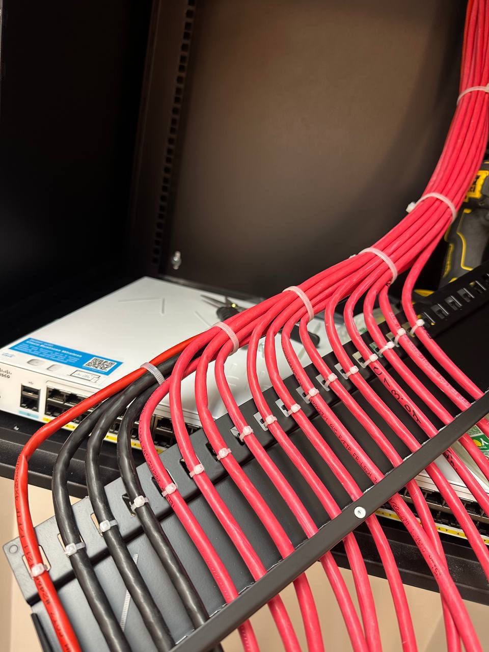 Zorganizowana wiązka czerwonych i czarnych kabli sieciowych w szafie rack, uporządkowanych za pomocą opasek zaciskowych, z widocznym switchem sieciowym Cisco oraz fragmentem narzędzia.