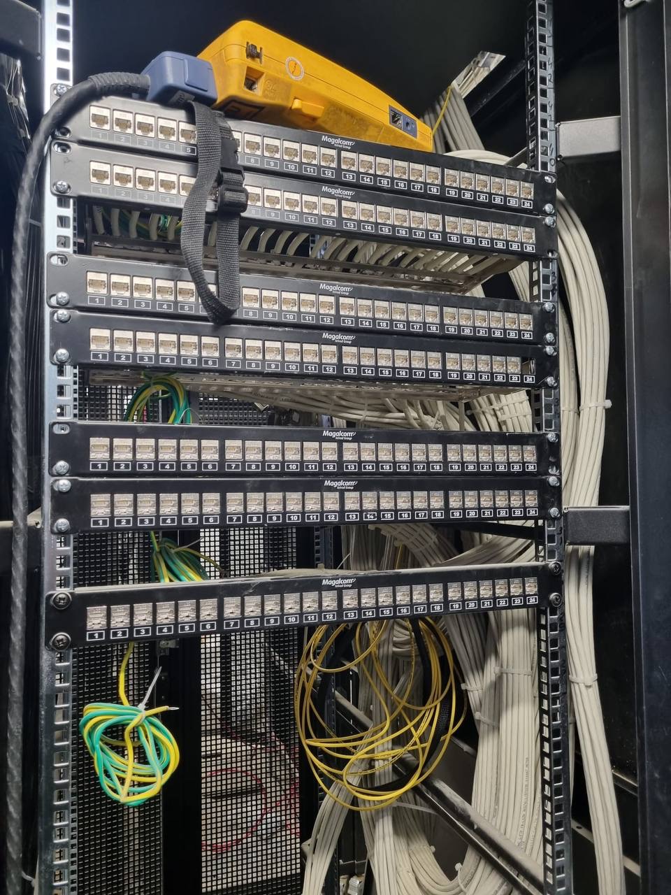 Patch panel w szafie rack z podłączonymi kablami Ethernet, żółty tester kabli na górze, widoczne kable ułożone w wiązki. Niektóre kable są podłączone, inne zwisają luźno.