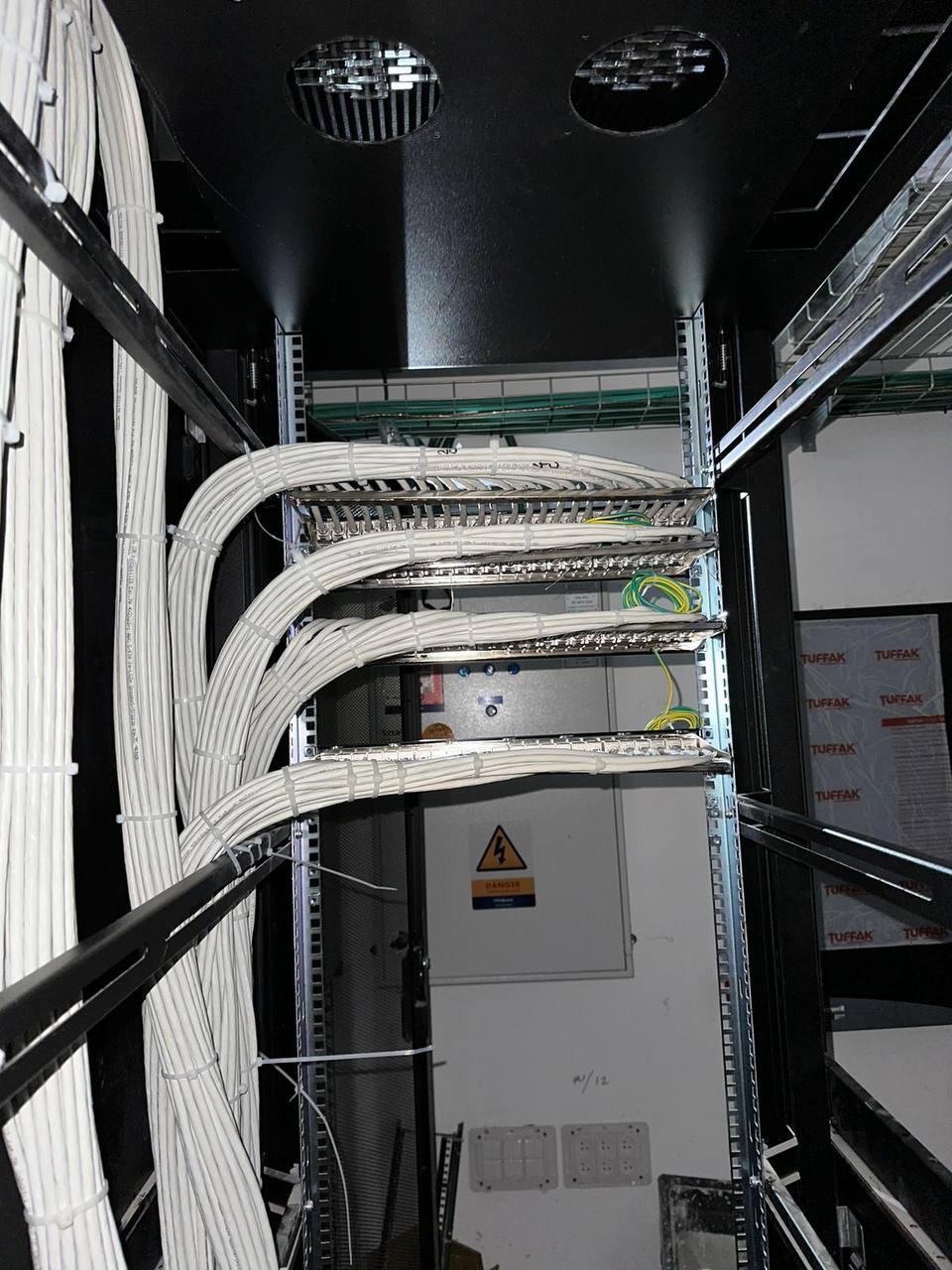 Widok z góry na uporządkowane kable sieciowe w szafie rack, zabezpieczone opaskami zaciskowymi, z widoczną skrzynką elektryczną z symbolem ostrzegawczym w tle.