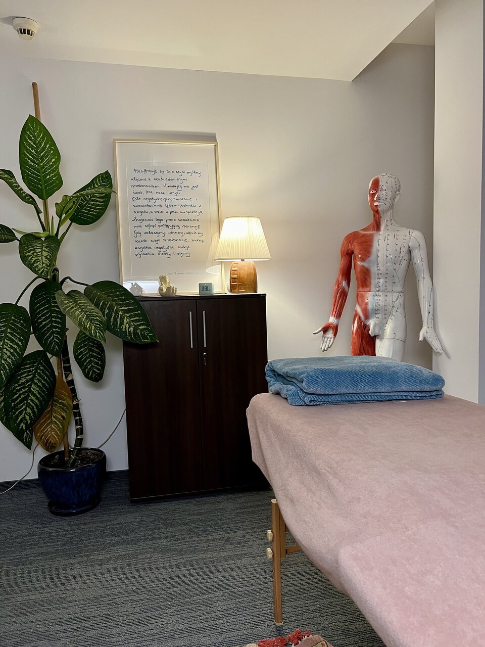 Gabinet z kozetką przykrytą różowym materiałem i niebieskim ręcznikiem, obok stoi model anatomiczny człowieka z zaznaczonymi punktami akupunkturowymi oraz szafka z lampką i ramką z tekstem.