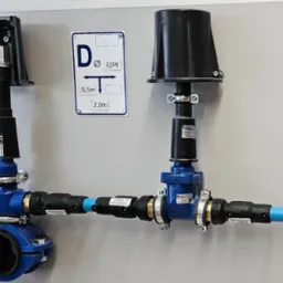Zestaw zaworów i rur z tworzywa sztucznego w kolorze niebieskim i czarnym, zamontowany na szarej ścianie, z widoczną tabliczką informacyjną o średnicy rury i odległościach.