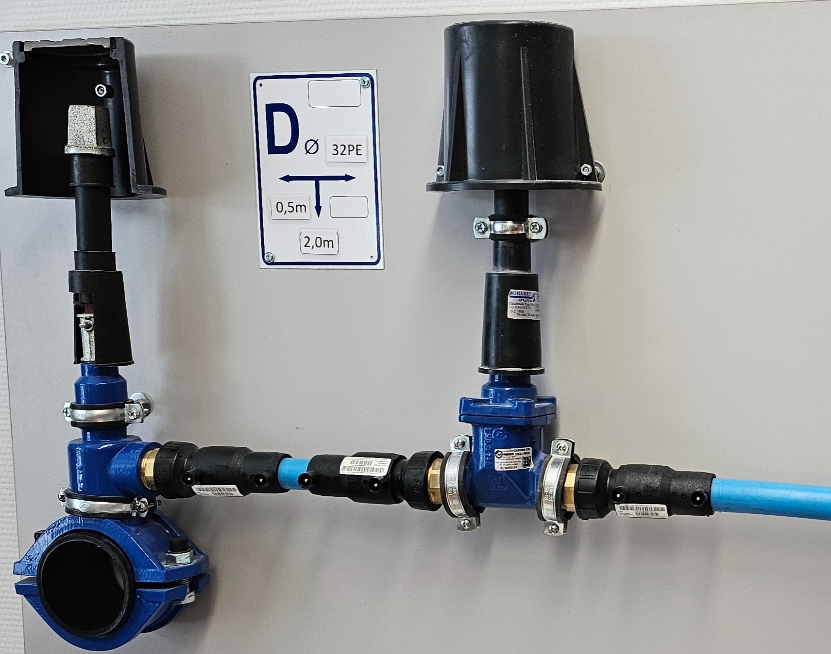 Zestaw zaworów i rur z tworzywa sztucznego w kolorze niebieskim i czarnym, zamontowany na szarej ścianie, z widoczną tabliczką informacyjną o średnicy rury i odległościach.