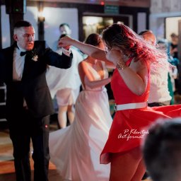 Piotr Klentak - Goście weselni tańczą podczas przyjęcia, panna młoda w białej sukni w tle, kobieta w czerwonej sukience z białym paskiem na pierwszym planie.